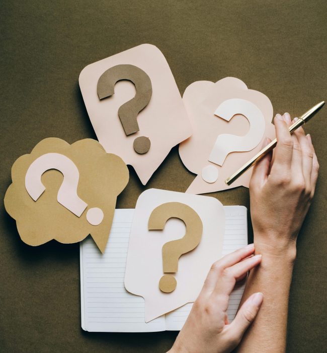 Hands holding pen next to paper question marks on notebook, symbolizing inquiry and creativity.